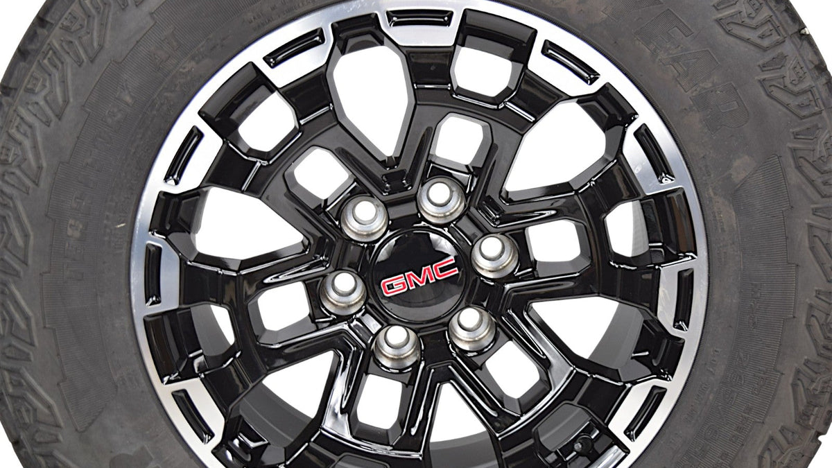 A close-up of a black and silver multi-spoke alloy wheel with a red GMC logo in the center, mounted on a black tire against a white background.