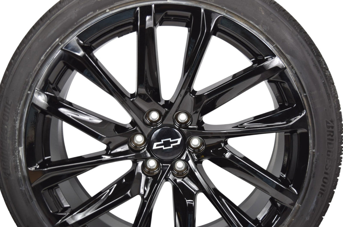 A close-up of a glossy black multi-spoke alloy wheel rim with a center cap featuring the Chevrolet logo, mounted on a black tire. The background is solid white.