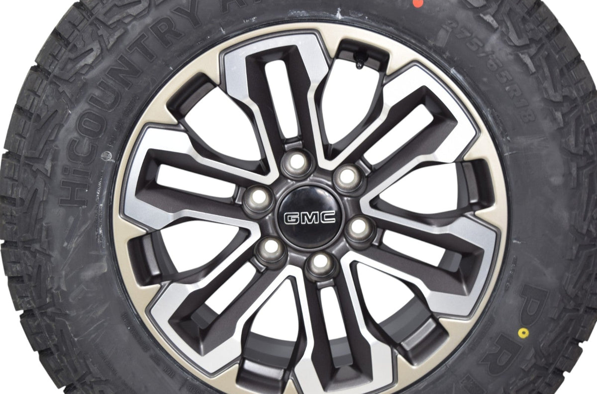A close-up of a black off-road tire mounted on a multi-spoke GMC truck wheel, shown against a plain white background.